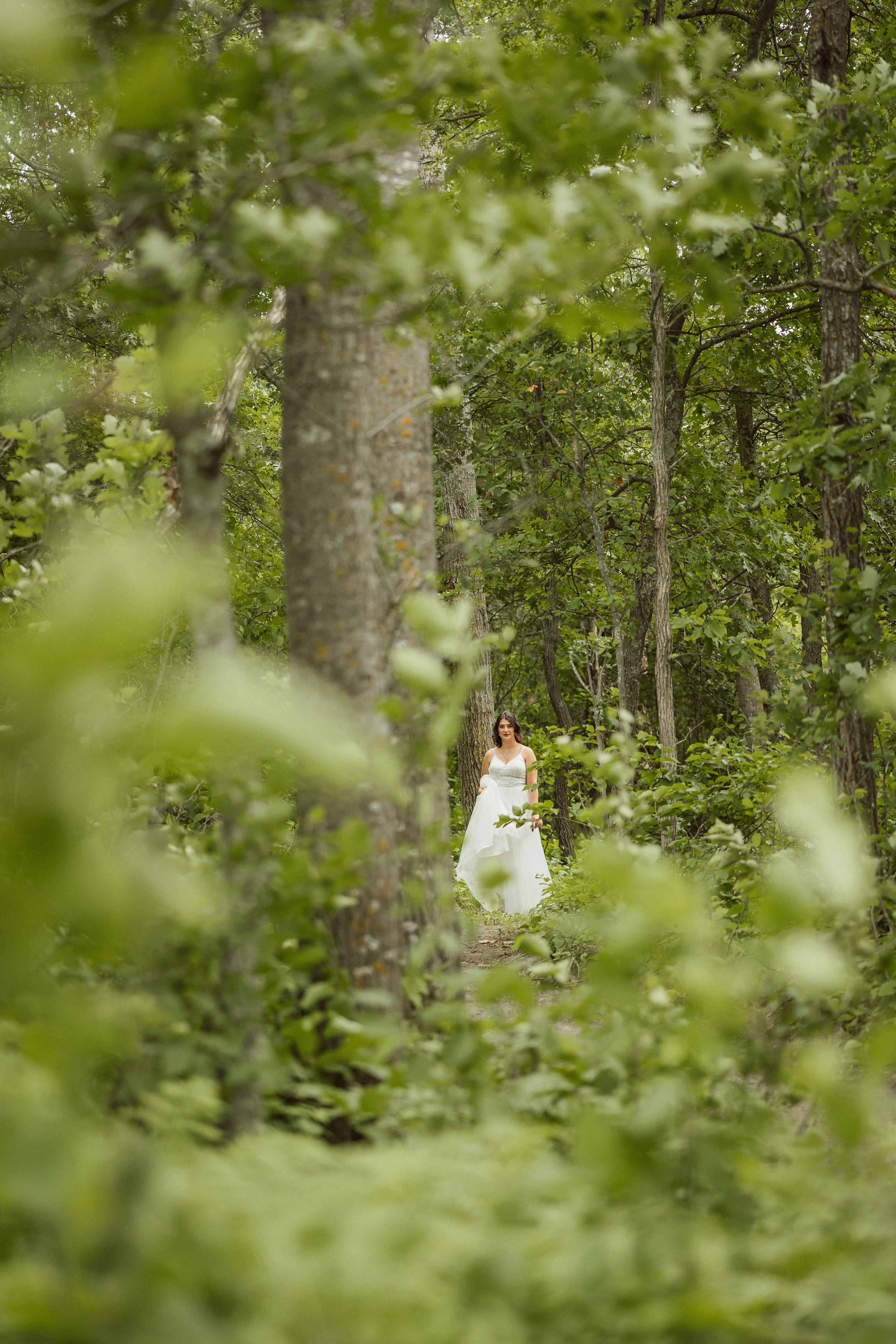 A bride in a white gown walking a forest path framed by lush green foliage