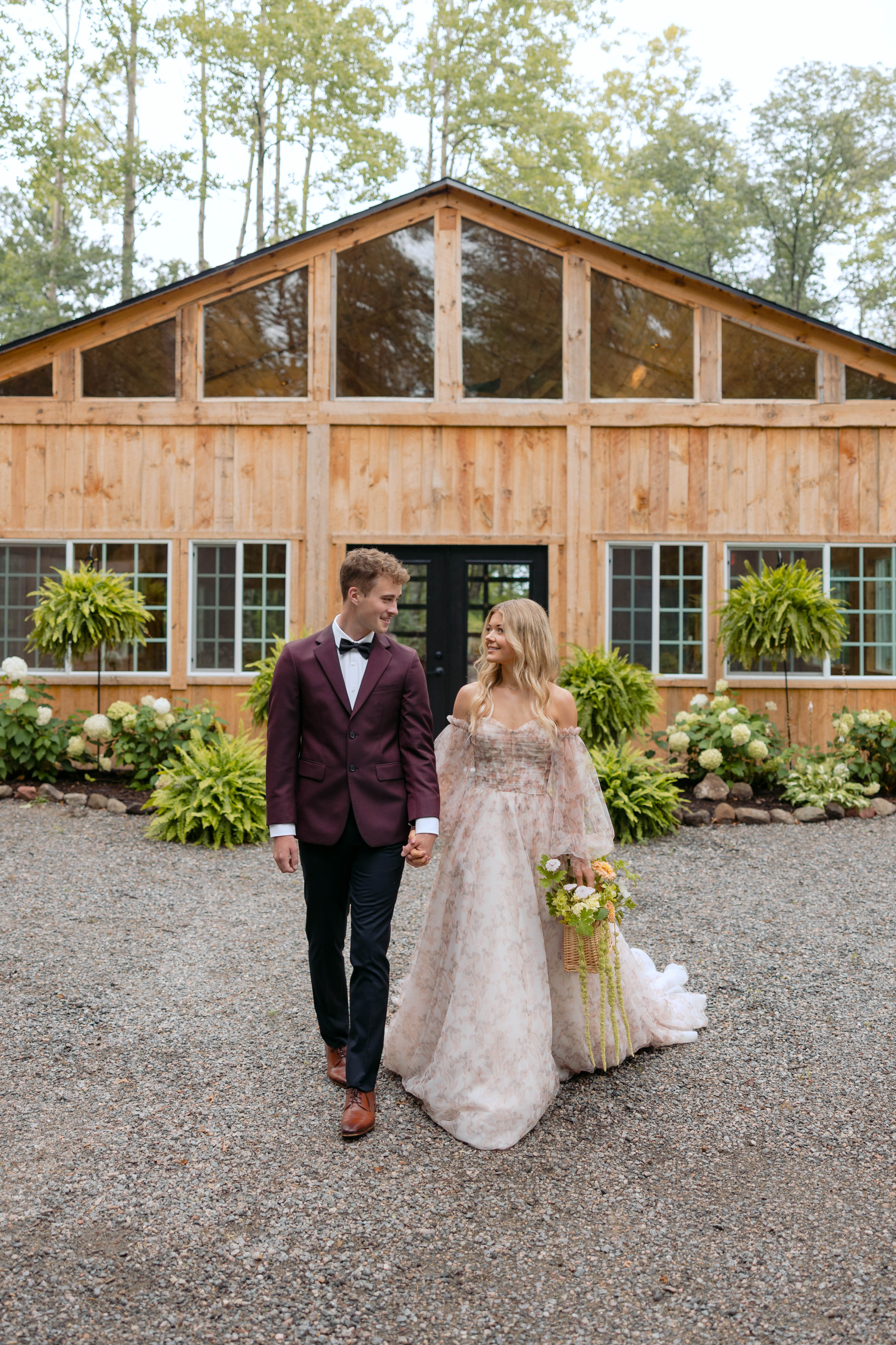 A couple walking hand in hand in front of the timber Emerald Cottage with its glass gable, hanging ferns, and white hydrangeas