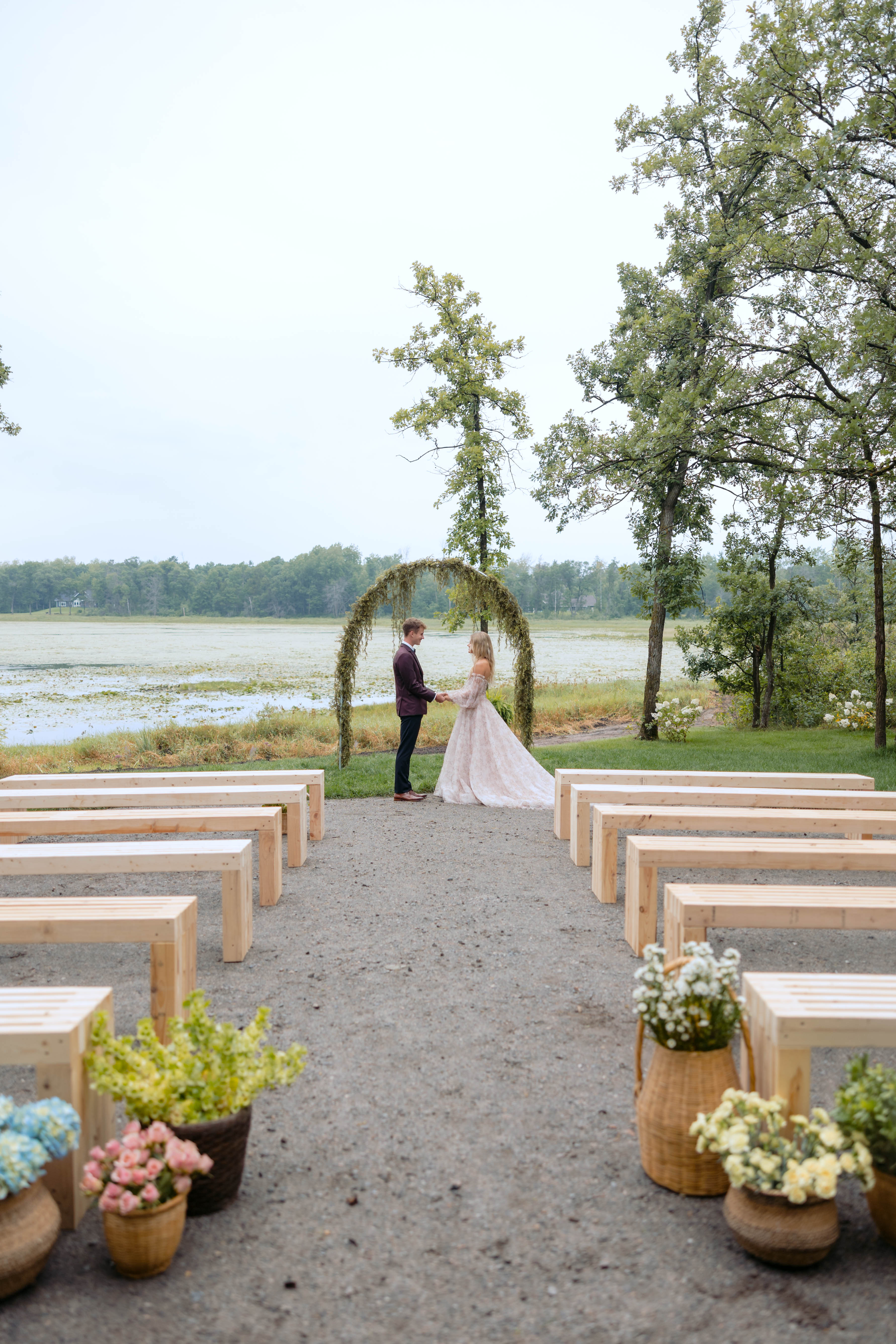 A couple holding hands beneath a greenery arch overlooking the lily-pad lake, with wooden benches and flower baskets lining the aisle