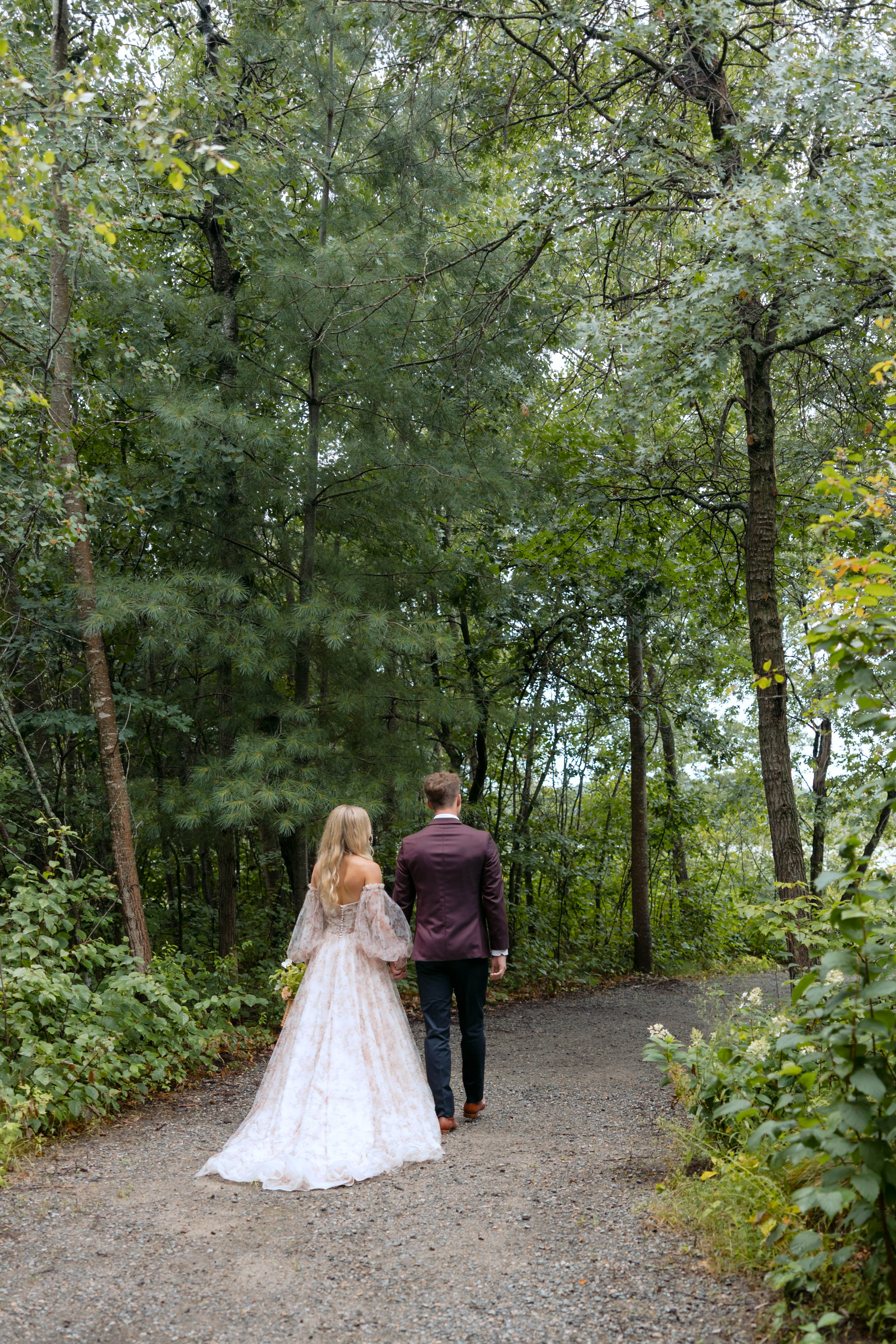 A couple walking hand in hand down a gravel path through the lush green woods