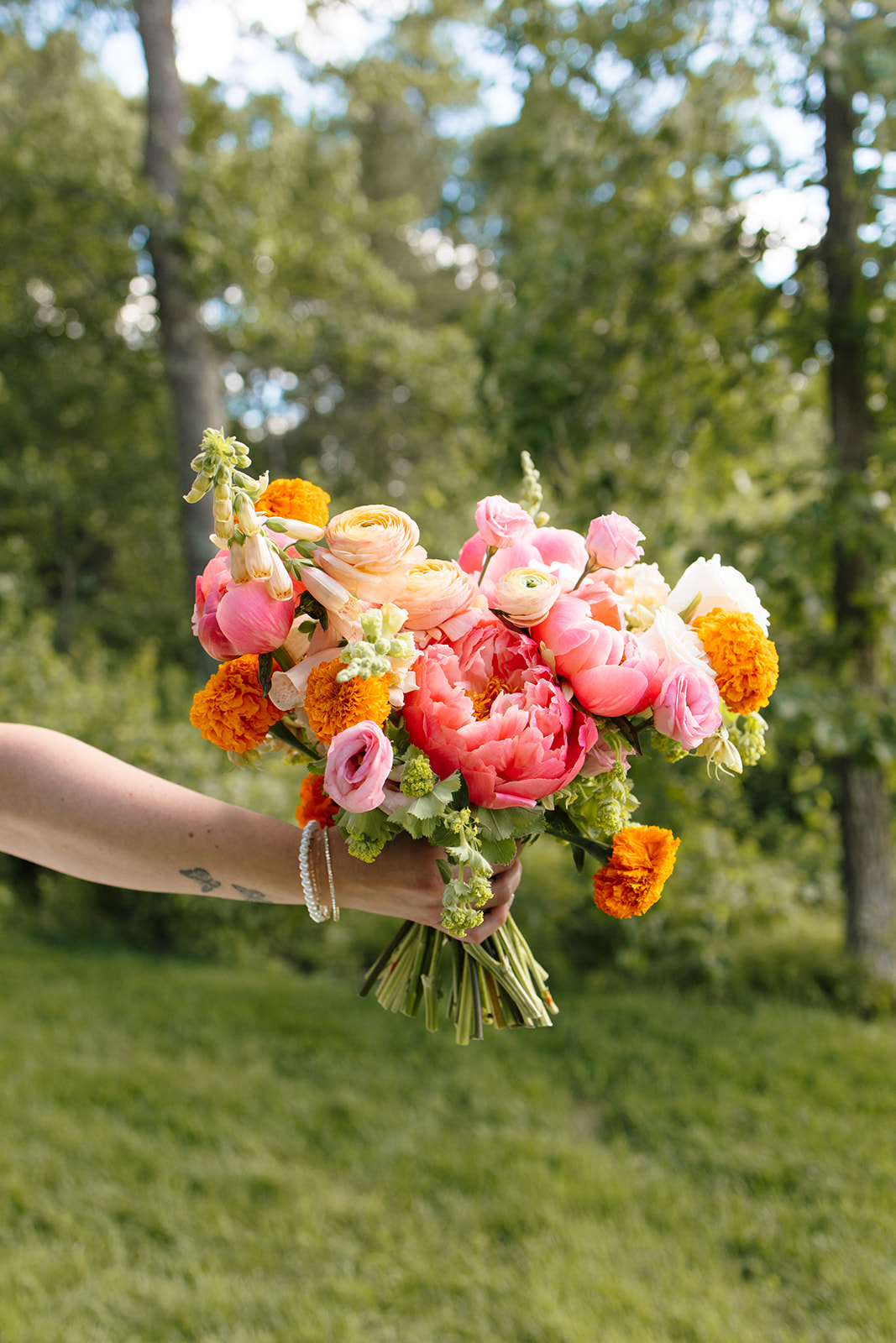 A colorful bridal bouquet of coral, peach, pink, orange, and white flowers held in the cottage woods