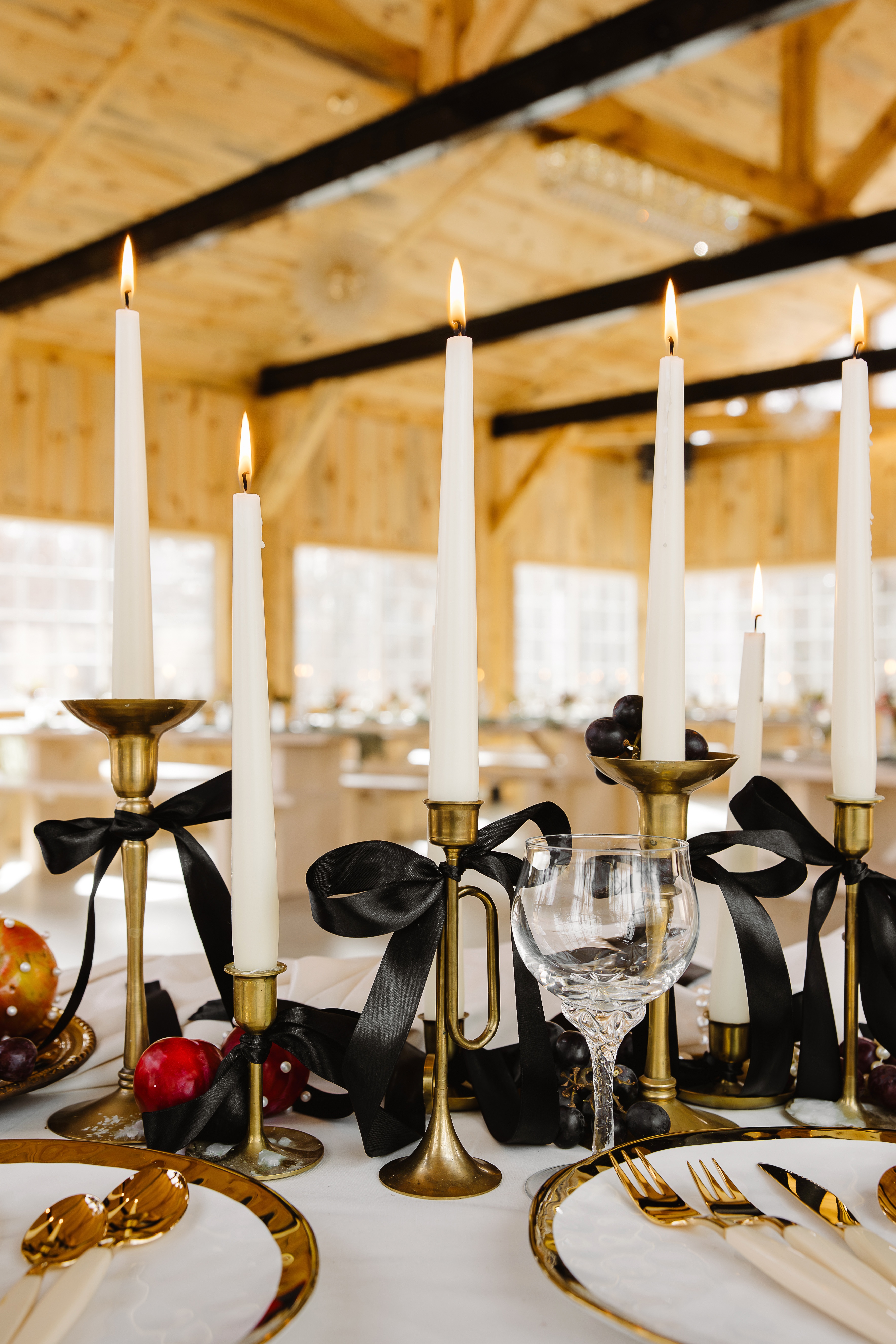 A candlelit dinner table with brass holders, black ribbon, grapes, and glassware