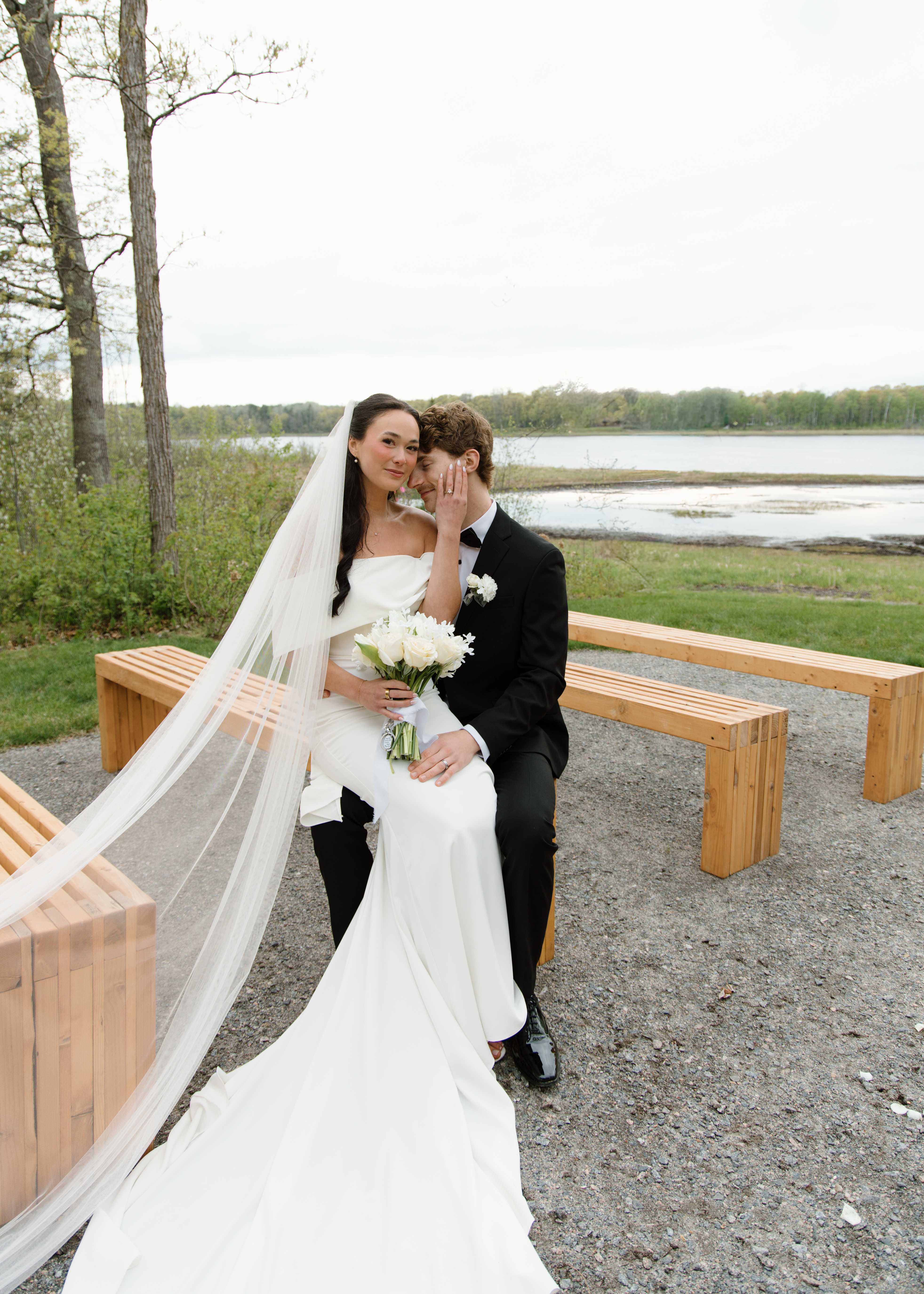 A couple exchanging vows beneath the greenery-covered lakeside arch with the officiant, guests, and lake behind them