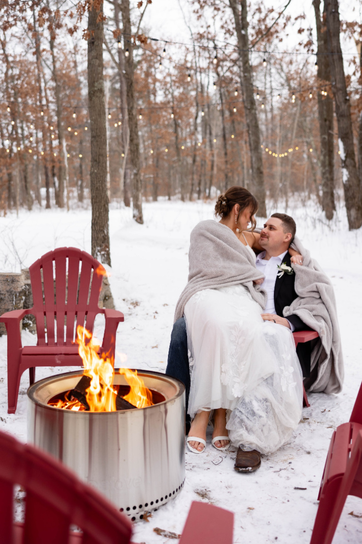 A winter couple wrapped in blankets beside a fire pit under outdoor string lights