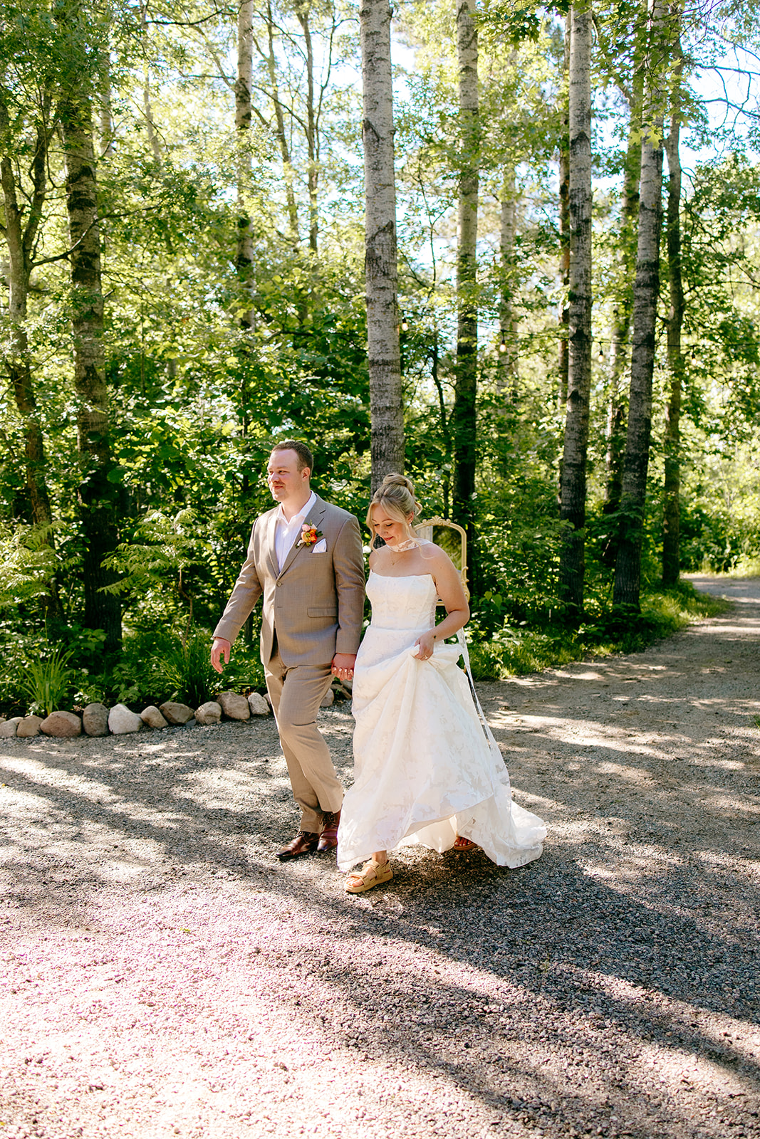 A couple walking hand in hand on the sunlit birch-lined path at the Emerald Cottage, photographed by Madisen Watson
